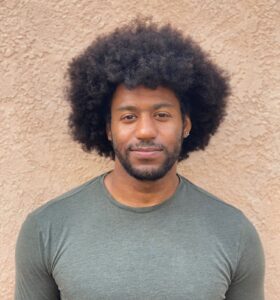 Kyle, a Black masculine presenting person with an afro hairstyle and close-cropped facial hair, stands in front of a stucco-textured wall in a simple gray shirt